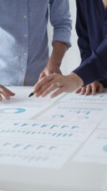 İş arkadaşları mali raporları inceliyor ve masadaki çizelge ve grafiklere işaret ediyor. Verileri analiz etmek ve stratejileri planlamak için birlikte çalışıyorlar.