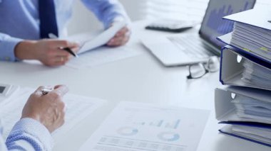 Ofis çalışanları mali raporları analiz ediyor ve ön planda evrak dolu mavi dosyalarla dolu beyaz bir masada otururken belgeleri imzalıyorlar. İş dünyasında denetim ve vergi teması