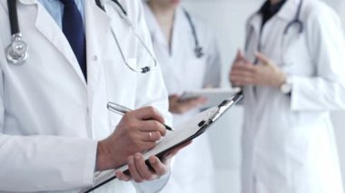 Steteskoplu beyaz önlüklü doktor hasta kaydını panoya yazarken, meslektaşlar arka plandaki davaları, takım çalışmasını ve profesyonel klinik danışmanlığı temsil eden davaları tartışıyorlar.