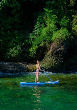 SUP, ayağa kalk. Taş kayaların yanında yeşil denizde yelken açan mayolu sarışın kız. Yaz tatilindeki kadın yaşam tarzı. Mtsvane Kontskhi Plajı, Batumi, Georgia.