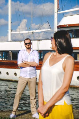 Limanda yürüyen mutlu aşıklar çifti. Gemi arka planda. Neşeli, beyaz elbiseli, gülümseyen adam ve beyaz bluzlu ve sarı pantolonlu şanslı kadın, açık deniz limanında..