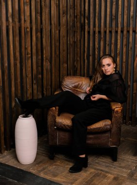 Siyah bluz ve topuklu ayakkabılarla poz veren zarif sarışın kadın. Büyük deri sandalyede oturan şık bir kadın. Arkaplanda tahta duvar var..