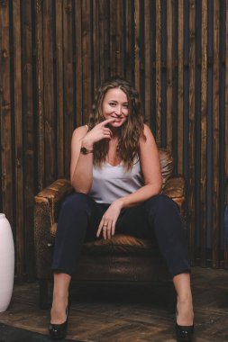 Beyaz bluz ve topuklu ayakkabıyla poz veren zarif sarışın kadın. Büyük deri sandalyede oturan şık bir kadın. Arkaplanda tahta duvar var..