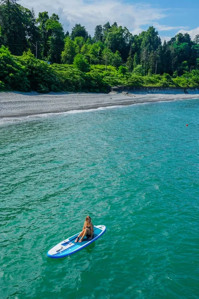 SUP, ayağa kalk. Sarışın kız denizde kürek çekerken dinleniyor. Hava görüntüsü. Arka planda Stone Beach. Yaz tatilindeki kadın yaşam tarzı. Mtsvane Kontskhi Plajı, Batumi, Georgia.
