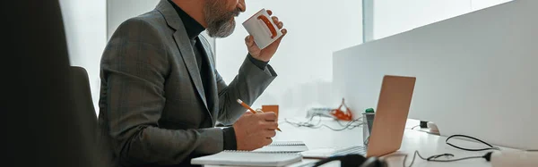 Side view of busy man sitting in modern office while drinking coffee and making notes on documents