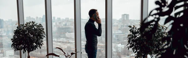 Busy man standing near panoramic window and talking on the phone