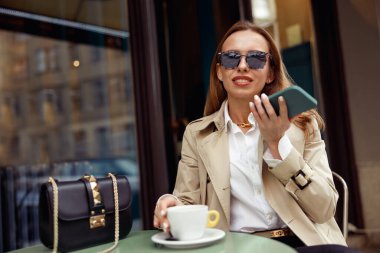 Woman recording audio message on mobile phone while drinking coffee on cafe terrace