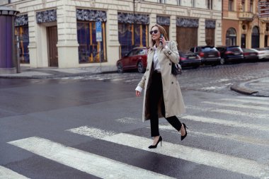 Woman in trendy outfits crosses road against backdrop of city buildings. High quality photo