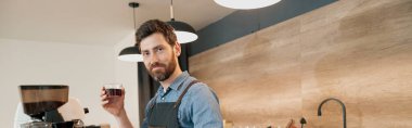 Smiling barista holding in hand glass with filter coffee and looking at the camera in coffee shop