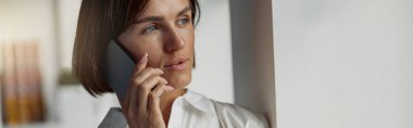 European business woman holding cup of coffee and talking phone while standing near window at office