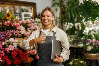 Çiçekçi dükkânı sahibi dizüstü bilgisayarlı çiçekçi çiçek arka planında baş parmak gösteriyor.