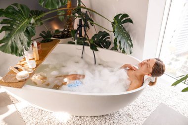 Evdeki modern beyaz baloncuk dolu küvetin üzerinde vücut bakım kozmetikleriyle yıkanan rahat genç bir kadın. Spa, sağlık konsepti