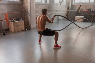 Savaş halatıyla spor salonunda egzersiz yapan güçlü, üstsüz, kaslı bir adamın arkası.