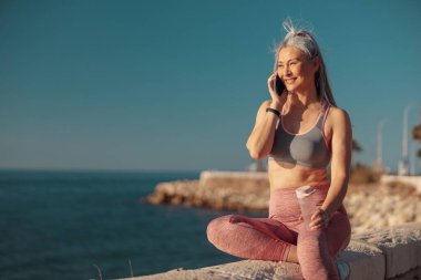 Spor elbiseli aktif bir kadın deniz kenarında oturuyor, cep telefonunu tutuyor ve konuşuyor, ileriye bakıyor.