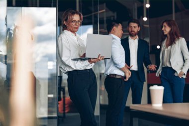 Mature confident businesswoman at office with group of colleagues on background, working on laptop. 