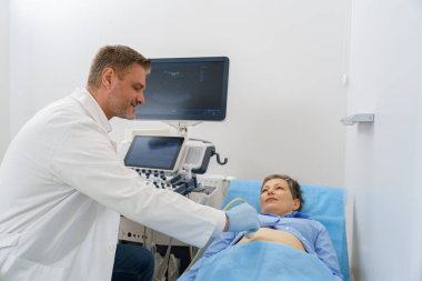 Orta yaşlı kadın hasta yatakta yatıyor ve klinikte ultrason muayenesi yaptırıyor.