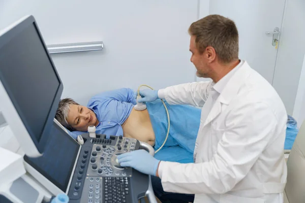 Middle aged woman patient lying on bed and having ultrasound examination of abdomen in clinic