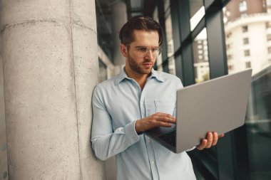 Laptop üzerinde çalışan yakışıklı girişimci iş günü boyunca modern iş birliği içinde duruyor.