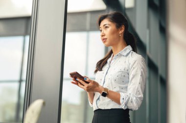 Modern ofis arka planında akıllı telefonu elinde tutarken yan bakan kadın girişimci 