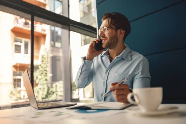 İşadamı iş yerinde çalışırken ve dizüstü bilgisayar üzerinde çalışırken müşteriyle telefonda konuşuyor.