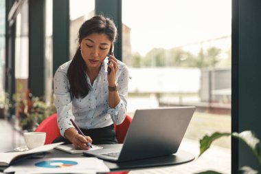 Kadın ofis müdürü müşteriyle telefonda konuşuyor ve ofiste otururken notlar alıyor. 