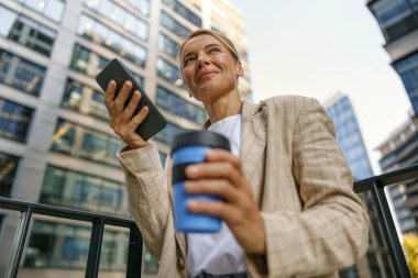 Gülümseyen iş kadını telefonu modern ofis terasında dururken kahve içer. 