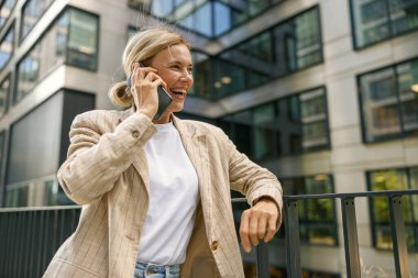 İş kadını telefonda konuşurken modern binanın arka planında dikilip yan tarafa bakıyor.