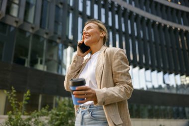 Kahve fincanı ile gülümseyen iş kadını modern binanın arka planında telefonla konuşuyor. 