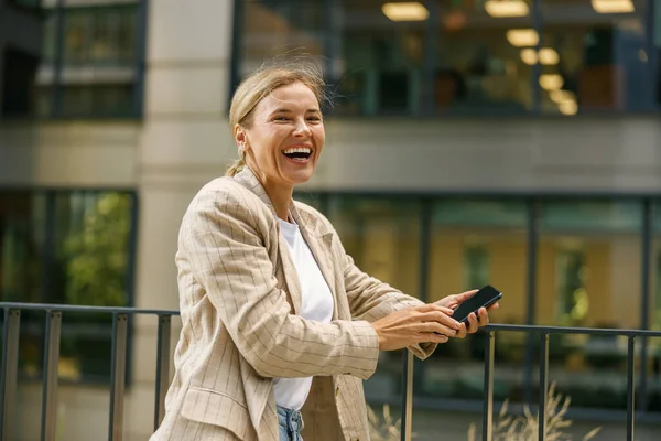 Smiling business woman holding phone while standing on modern office terrace and looks away