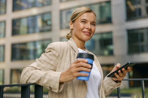 Smiling business woman drinks coffee while holding phone standing on modern office terrace