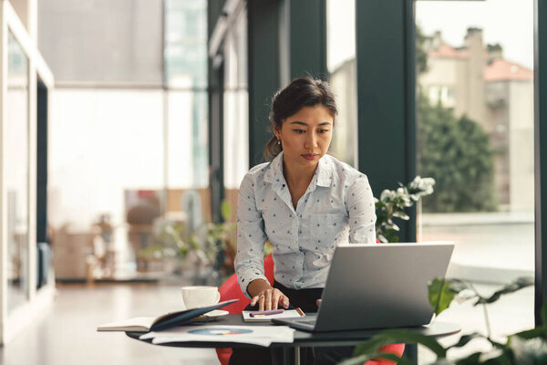 Focused stylish business woman working on laptop sitting the desk on modern office background 