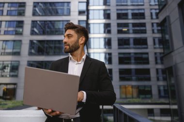 Diz üstü bilgisayarda çalışan şık bir erkek yönetici ofis terasında duruyor ve başka tarafa bakıyor. Yüksek kalite fotoğraf