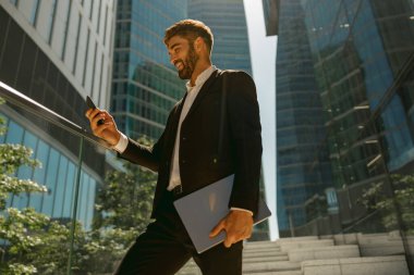 Gökdelenlerin arka planında dizüstü bilgisayarlı cep telefonu kullanan neşeli girişimci