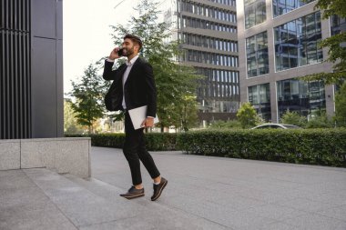 Takım elbiseli gülen girişimci modern binanın arka planında cep telefonuyla konuşuyor.