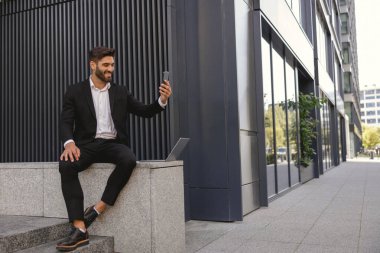 Gülümseyen işadamı cep telefonuyla özçekim yapıyor ofis binasının yanındaki bankta oturuyor.