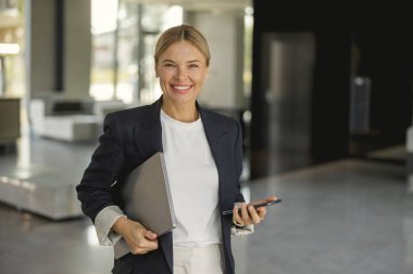 Modern ofis duvarının arka planında telefonuyla gülümseyen kadın girişimci kameraya bakıyor.