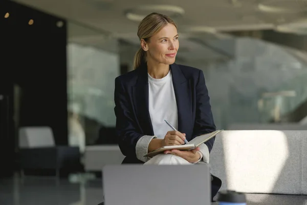 Pretty businesswoman making notes in note pad sitting on modern office background