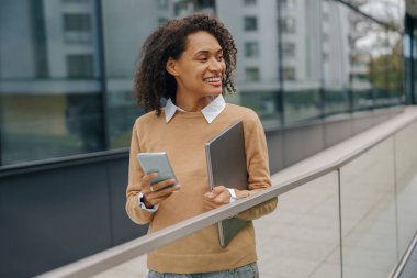 Gülümseyen kadın yönetici, modern bina arka planında dizüstü bilgisayarla dururken telefonu kullanıyor