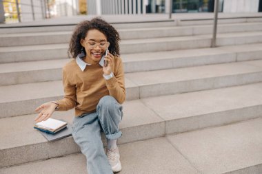 Keyifli iş kadını, binanın arka planında merdivenlerde otururken müşteriyle telefonda konuşuyor.