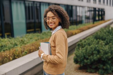 Gülümseyen kadın serbest yazar elinde not defterleriyle modern bina arka planında duruyor.