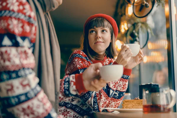 In a cozy cafe setting, a woman in a vibrant Christmas sweater cradles a cup of tea, embodying the warmth of the season