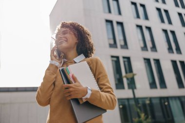 Güzel bayan girişimci modern bina arka planında dizüstü bilgisayarla konuşurken telefonla konuşuyor.