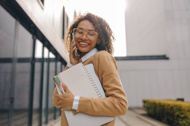 Gülümseyen kadın yönetici modern bina arka planında dizüstü bilgisayarla konuşurken telefonla konuşuyor.