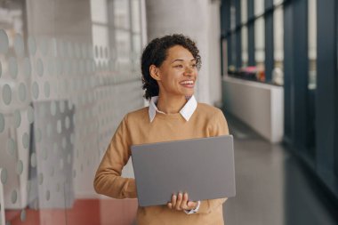 Genç gülümseyen iş kadını, modern ofis geçmişinde dururken dizüstü bilgisayar kullanıyor. 