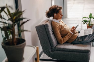 Kadın yönetici, pencerenin yanındaki modern ofiste dizüstü bilgisayarda çalışırken telefonu kullanıyor
