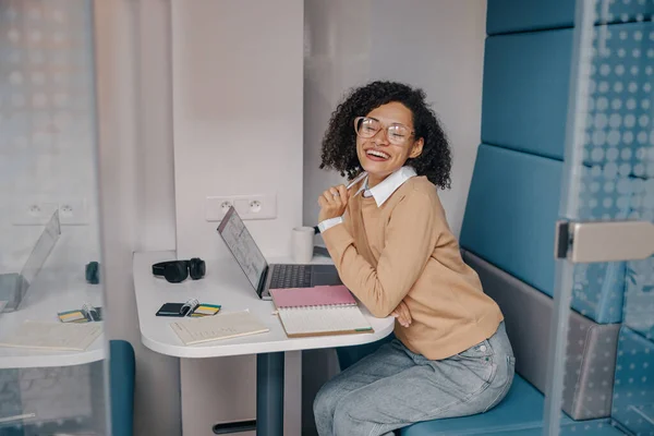 Smiling female analyst in eyeglasses working on laptop and making notes while sitting in cozy office