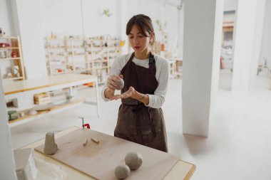 Genç bir kadın çömlek atölyesindeki ahşap bir masada kupa yapmak için kil hazırlıyor.