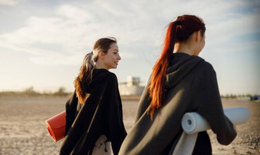 Sabah spor salonundan sonra iki zinde kadın yoga minderleriyle sahilde yürüyor. Yüksek kalite fotoğraf