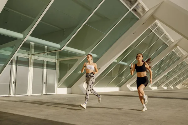 Spor giyimli iki atletik kadın sabahın erken saatlerinde şehirde koşuyor. Hedefe ulaşılıyor.