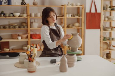 Çömlek atölyesindeki kullanılmamış kil vazo ile önlük giyen gülümseyen genç bayan seramistim.